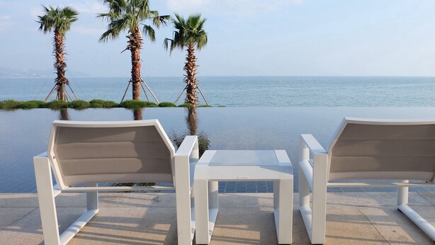 Dos sillones blancos y una pequeña mesa en una terraza con vista al mar y palmeras.