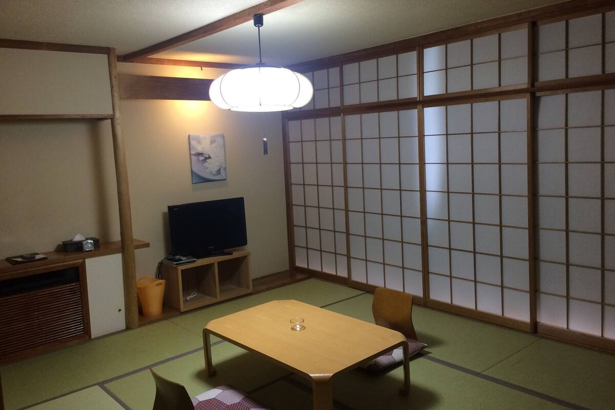 A traditional Japanese room with tatami flooring, a low table, two chairs, a television on a wooden stand, and a shoji screen in the background.