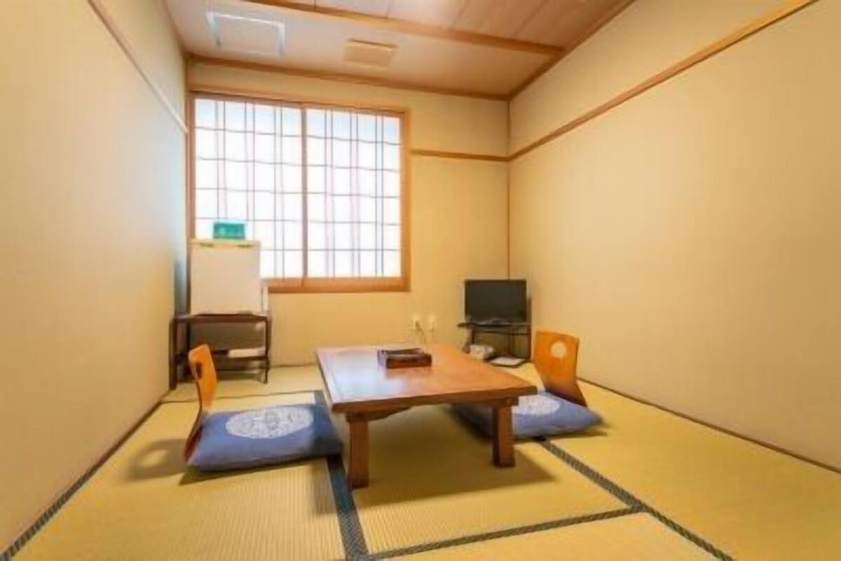Une pièce de style japonais traditionnel, avec un sol recouvert de tatamis, une table basse, deux coussins, un téléviseur et une fenêtre équipée de stores verticaux.