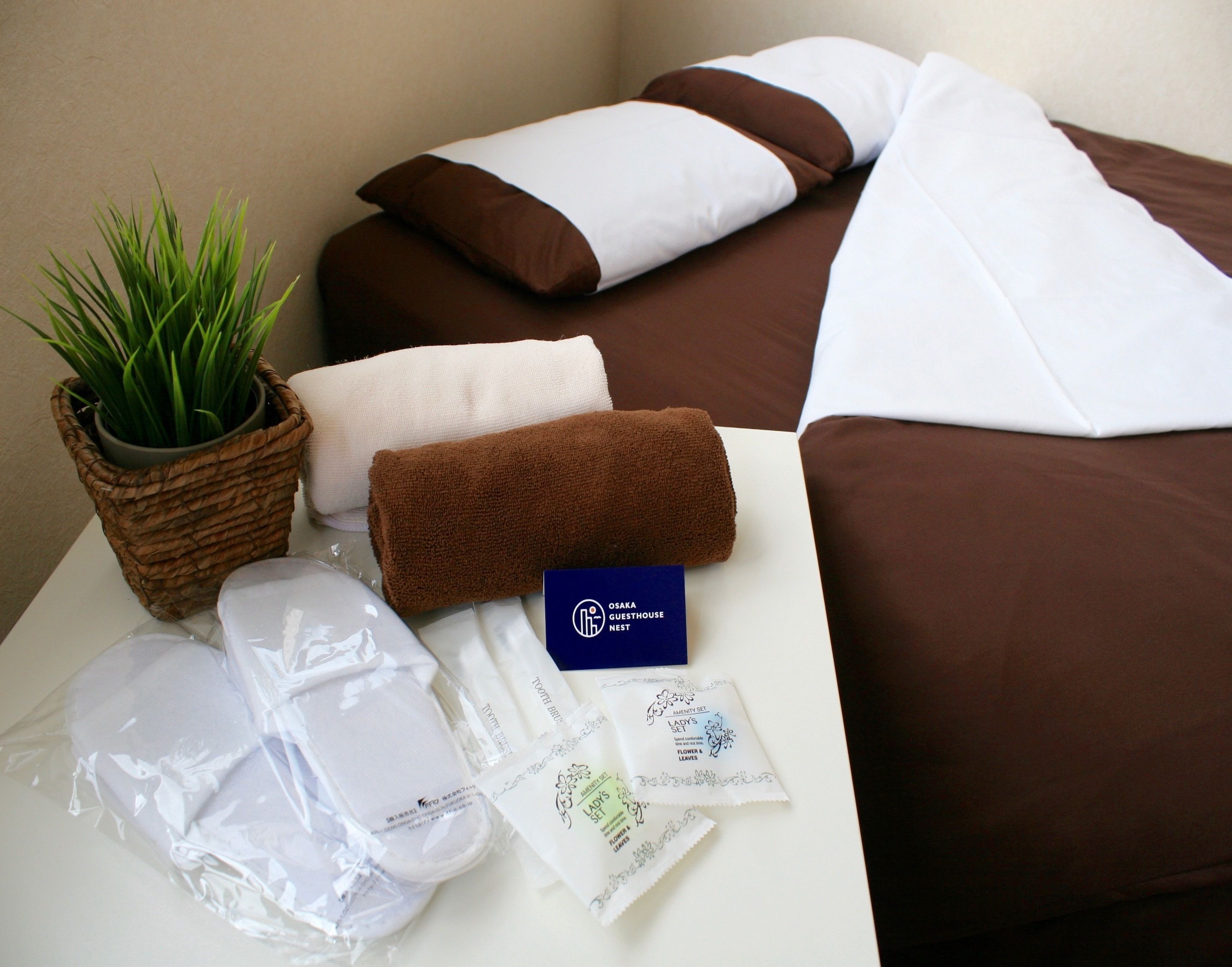 Ein ordentlich eingerichtetes Hotelzimmer mit Bett, Kissen, einem kleinen Tisch mit Toilettenartikeln und einer Zimmerpflanze.