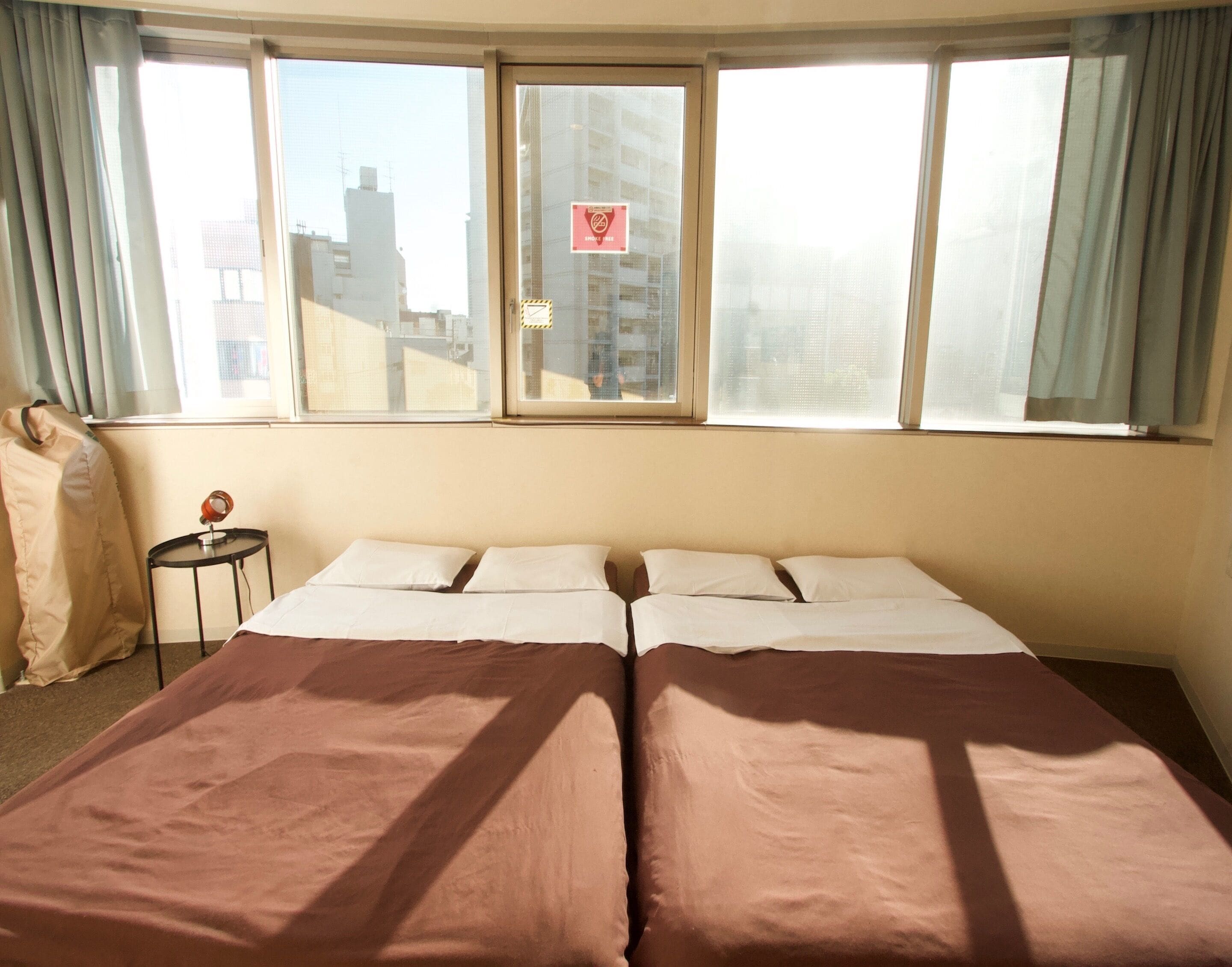 A twin bed room with a large window, a small table with a lamp, and a curtain on the left.