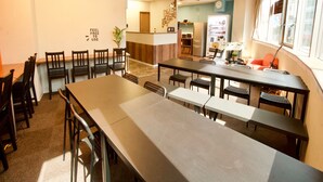 A spacious dining area with long tables and chairs, a kitchenette in the background, and a clock on the wall. - Osaka Guesthouse Nest - Hostel (Osaka)