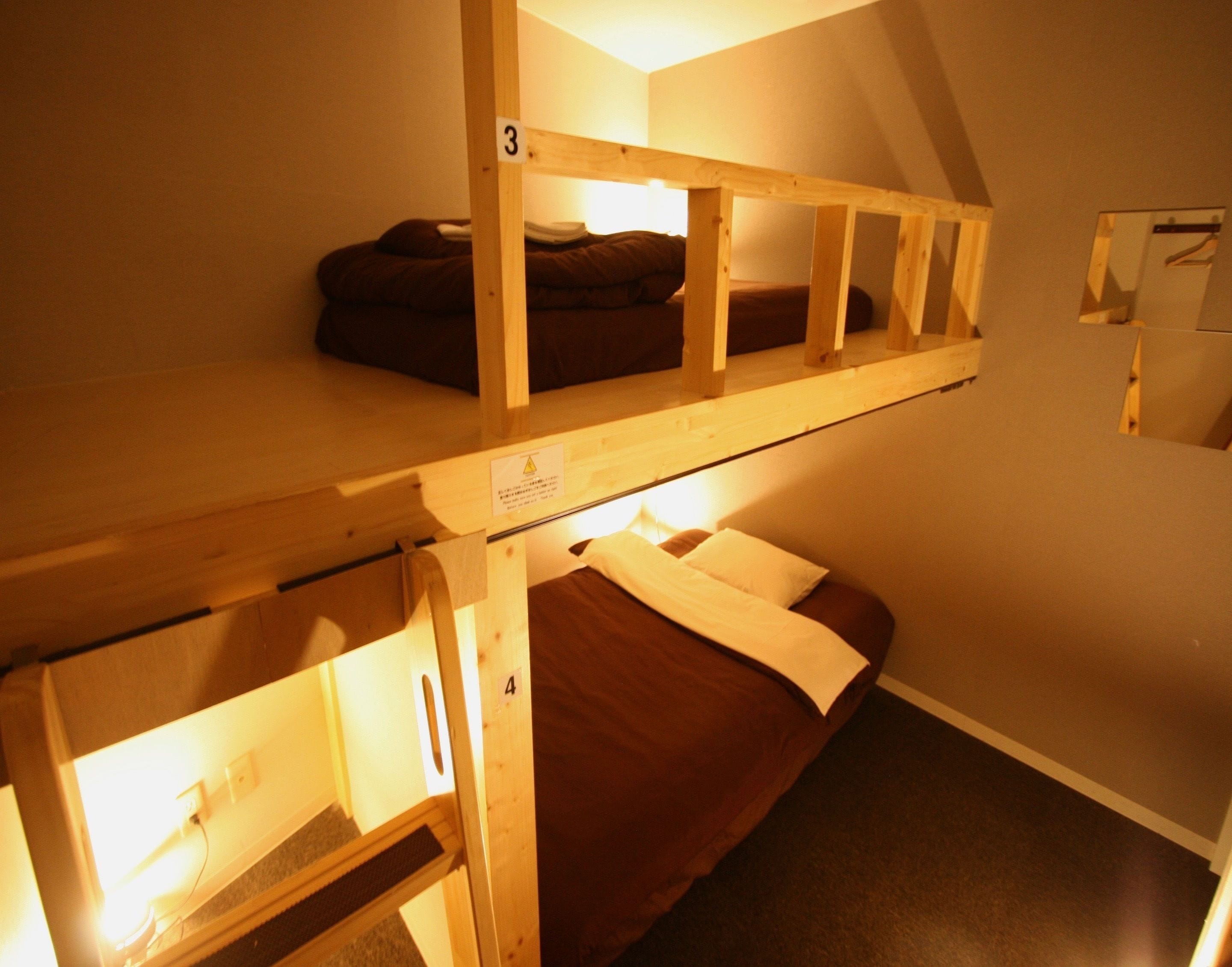 A dormitory-style room with two bunk beds, each with a mattress and pillows. There is a small shelf on the lower bunk and a wall-mounted shelf above the upper bunk.