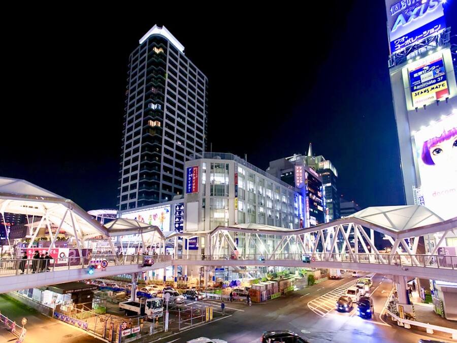 A modern urban area with a prominent high-rise building, a covered pedestrian walkway, and various commercial establishments.