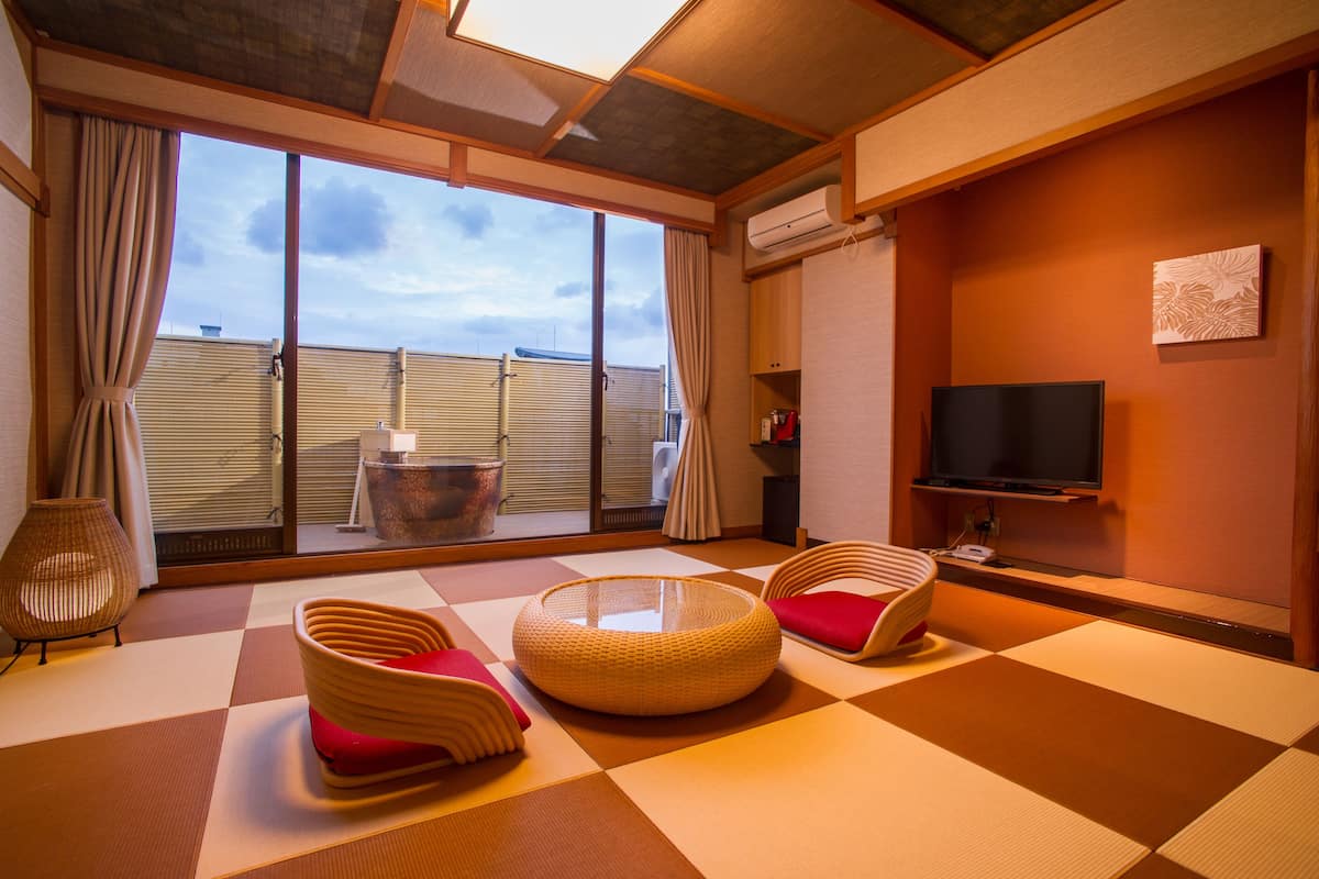 Una habitación con suelo de tatami, una mesa redonda de tatami, dos sillas de tatami, un televisor en la pared y vistas al exterior a través de amplias ventanas.