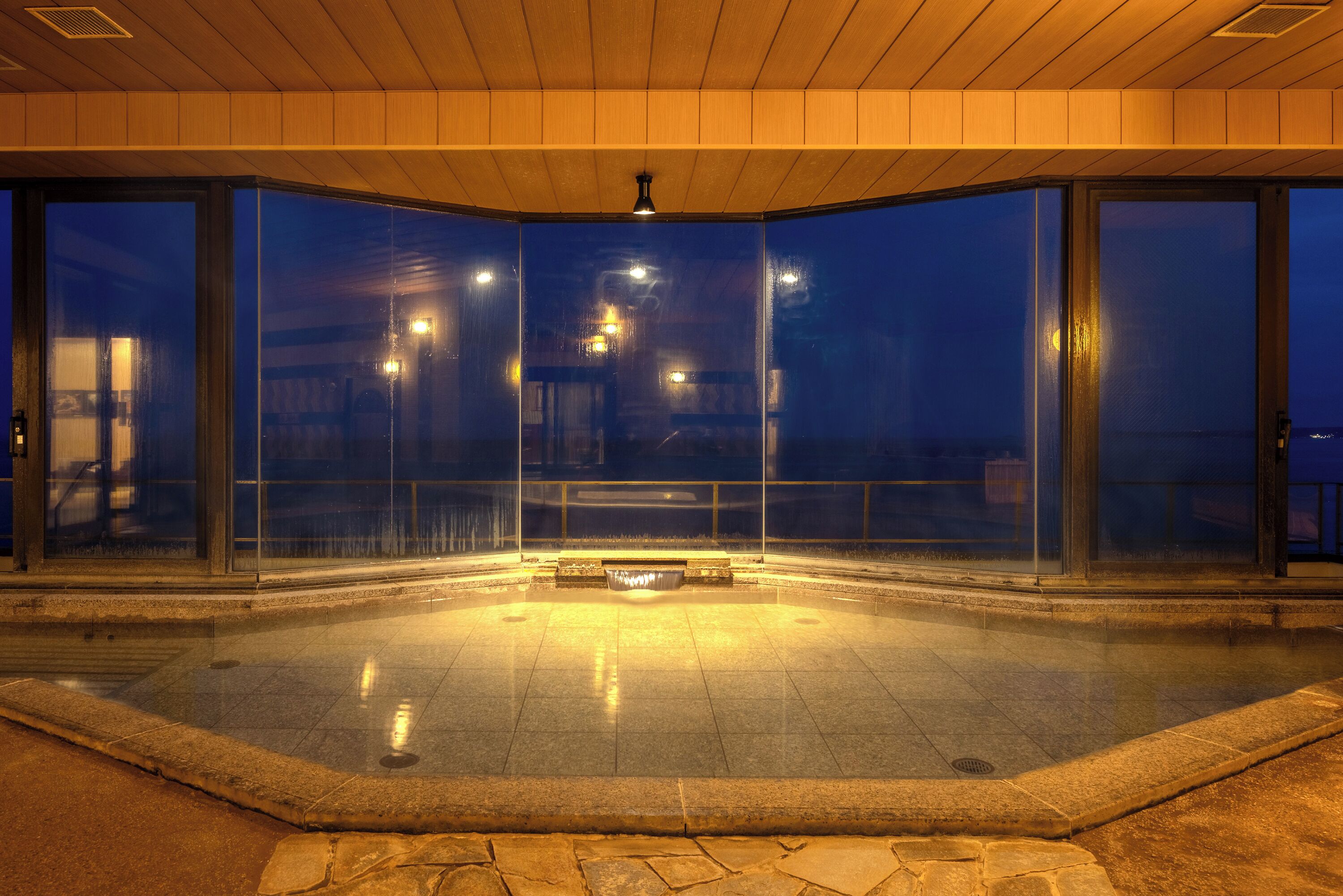 Un área de piscina interior con paredes de vidrio, un accesorio de luz central y un borde de piedra.