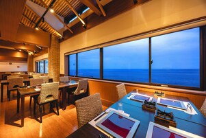 Uma sala de jantar com cadeiras e mesas de vime, grandes janelas com vista para o oceano e vigas de madeira no teto.