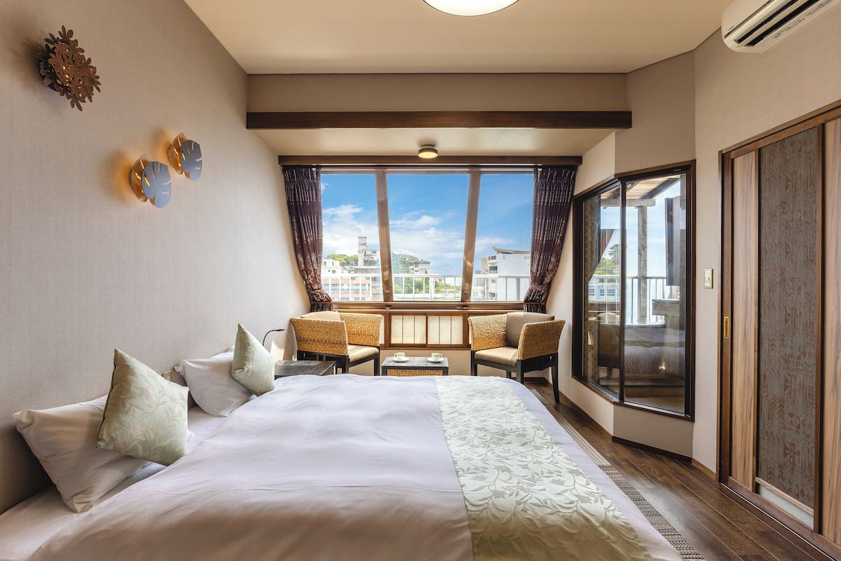 Habitación de hotel con una cama grande, una mesita pequeña, dos sillas, un ventanal con vistas a edificios y un armario.
