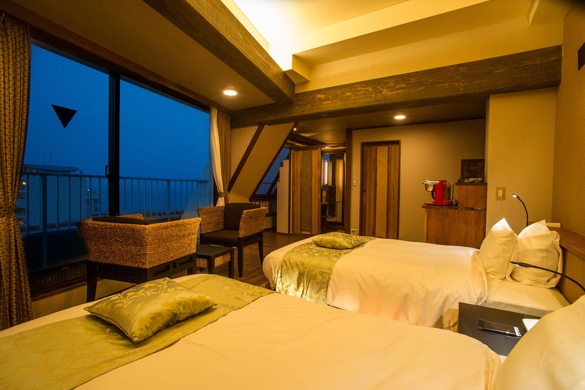 Habitación de hotel con dos camas, un escritorio, una silla y un balcón con vistas a la ciudad.