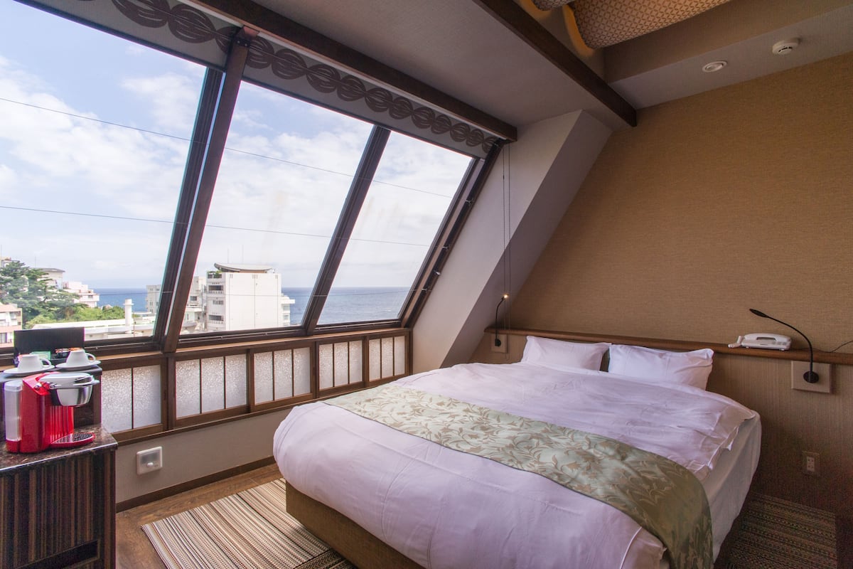 Habitación de hotel con un ventanal grande, una cama, una mesita de noche con un teléfono y vistas a la ciudad y al mar.