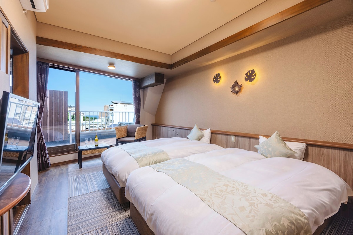 Habitación de hotel con dos camas, un ventanal grande, un televisor y vistas a la ciudad.