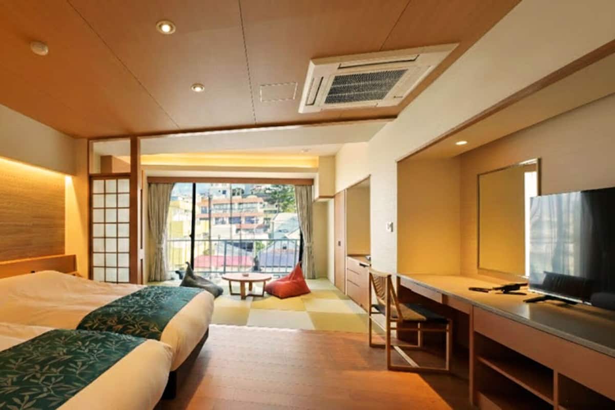 Habitación de hotel con dos camas, un escritorio con televisor y vistas a edificios.