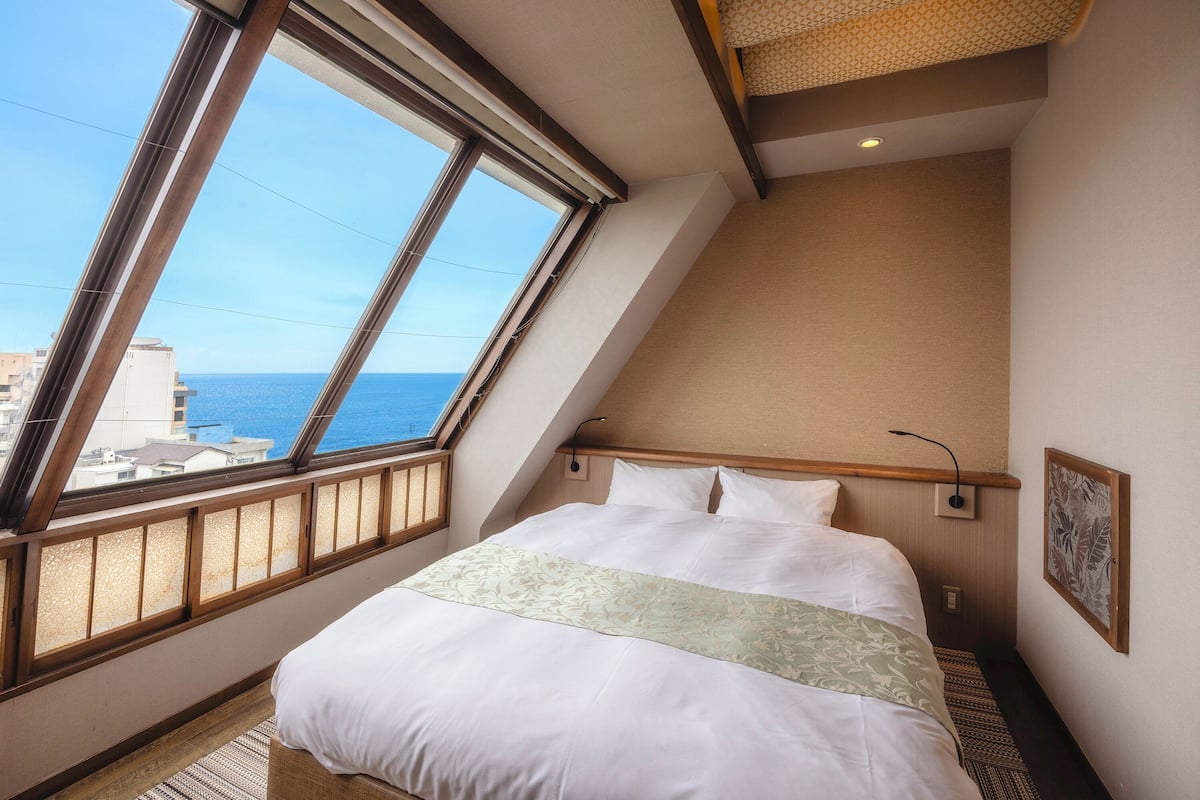 Un dormitorio con un ventanal grande, una cama con ropa blanca, un cabecero y una lámpara de pared.