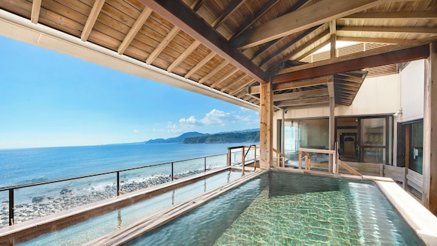A clear view of a swimming pool with a wooden deck and a glass barrier, overlooking a rocky shoreline and the sea.