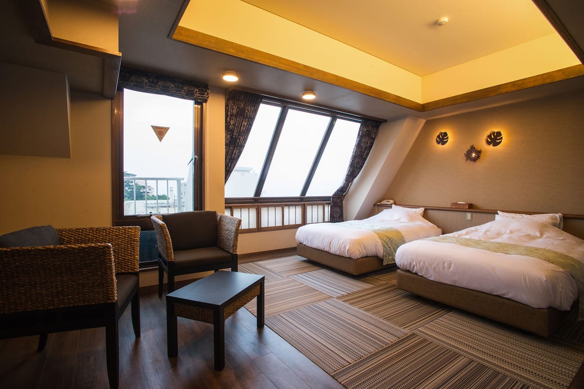 Habitación de hotel con dos camas, un sofá, una mesita y vistas a la ciudad a través de amplias ventanas.