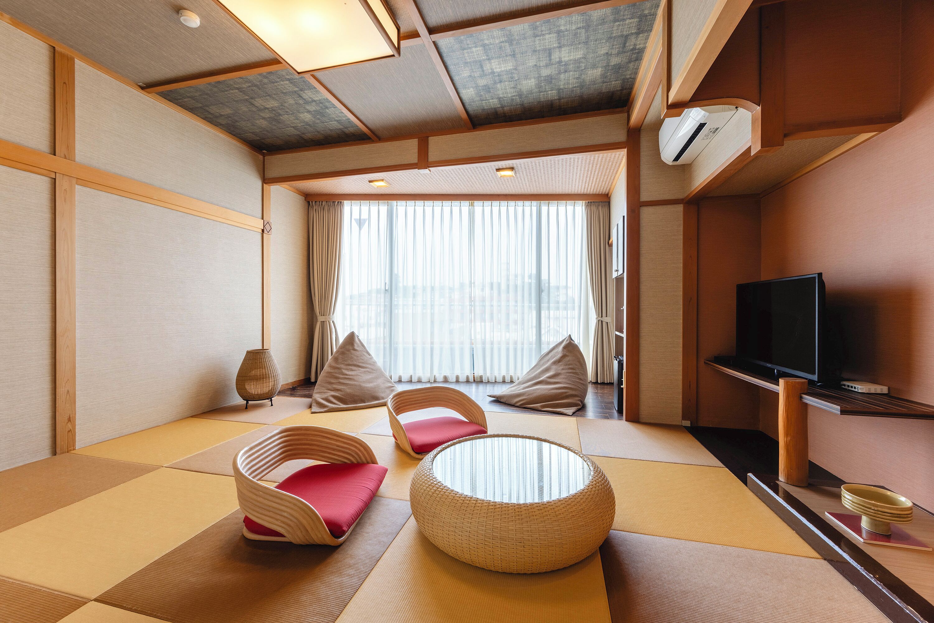 Une pièce de style japonais traditionnel, avec un sol recouvert de tatamis, une table basse, deux fauteuils rembourrés, un téléviseur et une grande fenêtre ornée de rideaux légers.