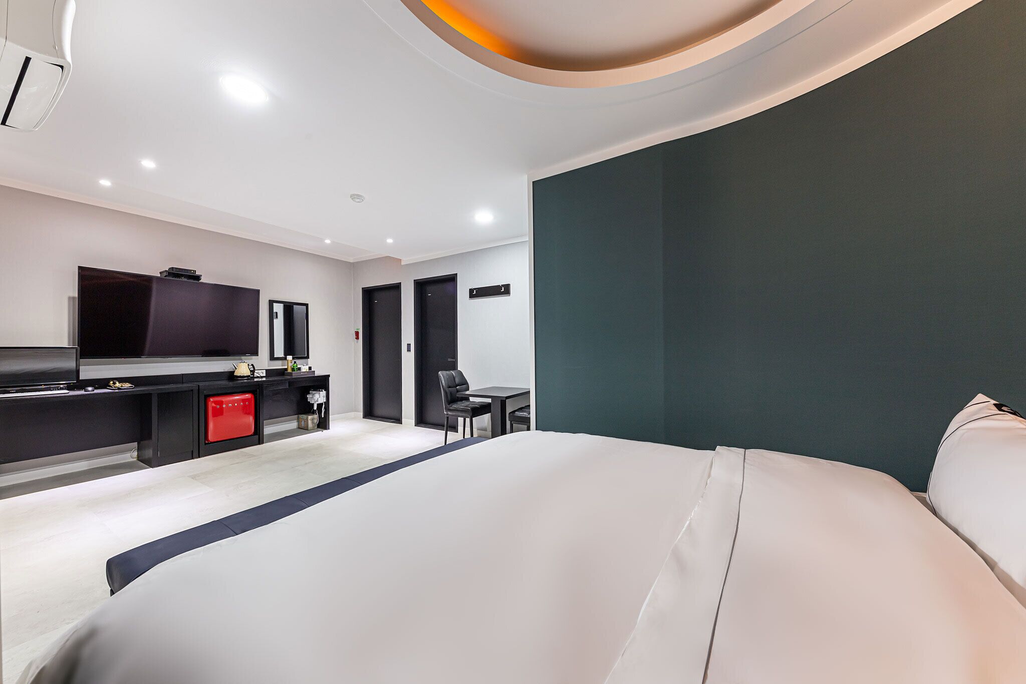 A modern hotel room with a large bed, a desk with a chair, a television, and a red minibar.