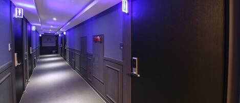 A hotel hallway with doors on both sides, emergency exit signs, and a fire alarm on the wall.