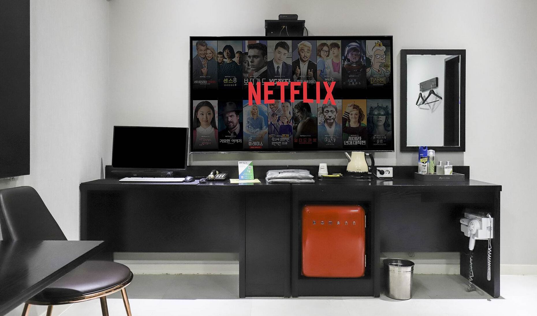 Una oficina moderna con un televisor de pantalla plana grande mostrando carteles de películas, un escritorio con computadora, teclado y ratón, un asiento acolchado rojo y un espejo en la pared.