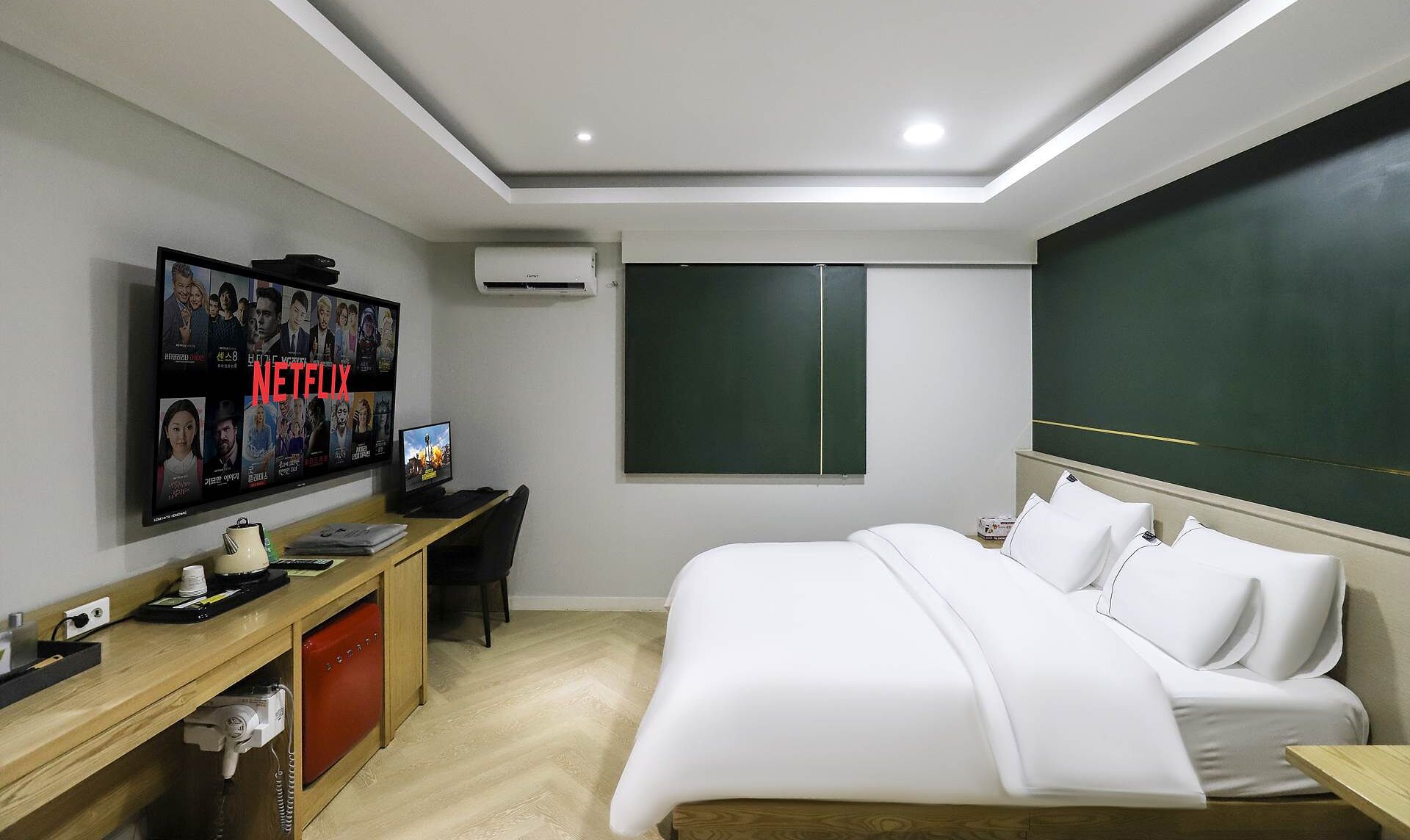 Una habitación de hotel moderna con una cama grande, un escritorio con computadora, un televisor montado en la pared y una pizarra verde.