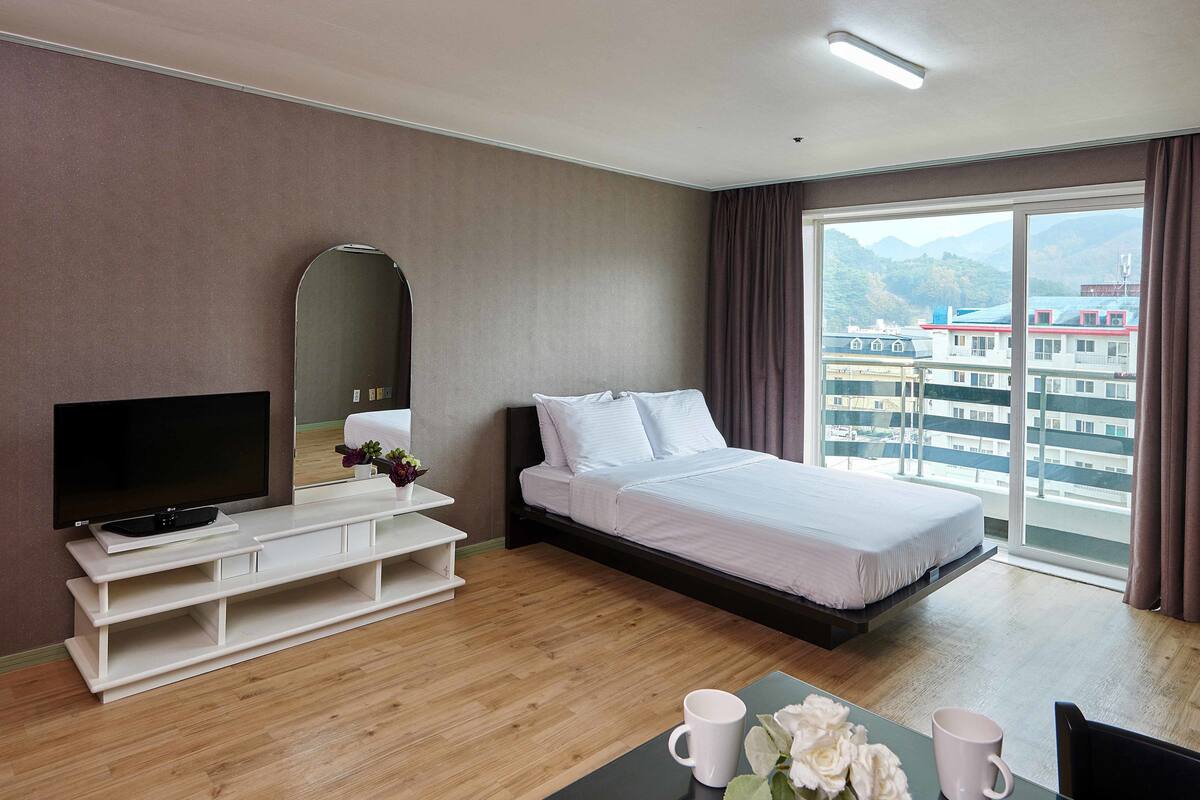 A hotel room with a bed, TV, and a view of buildings through a large window.