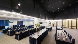 Un salón de conferencias con varias mesas y sillas dispuestas en filas, suelo alfombrado e iluminación empotrada en el techo.