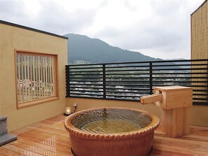 A wooden deck with a hot tub, a window with vertical bars, and a view of a mountain and cityscape. - Ryokan Biyunoyado (Yamanouchi)