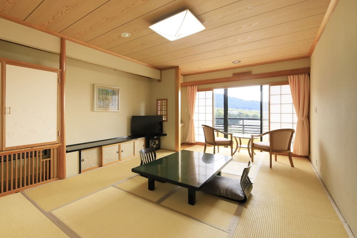 Una habitación japonesa tradicional con suelo de tatami, una mesa baja, sillas, un televisor y un ventanal con vistas.