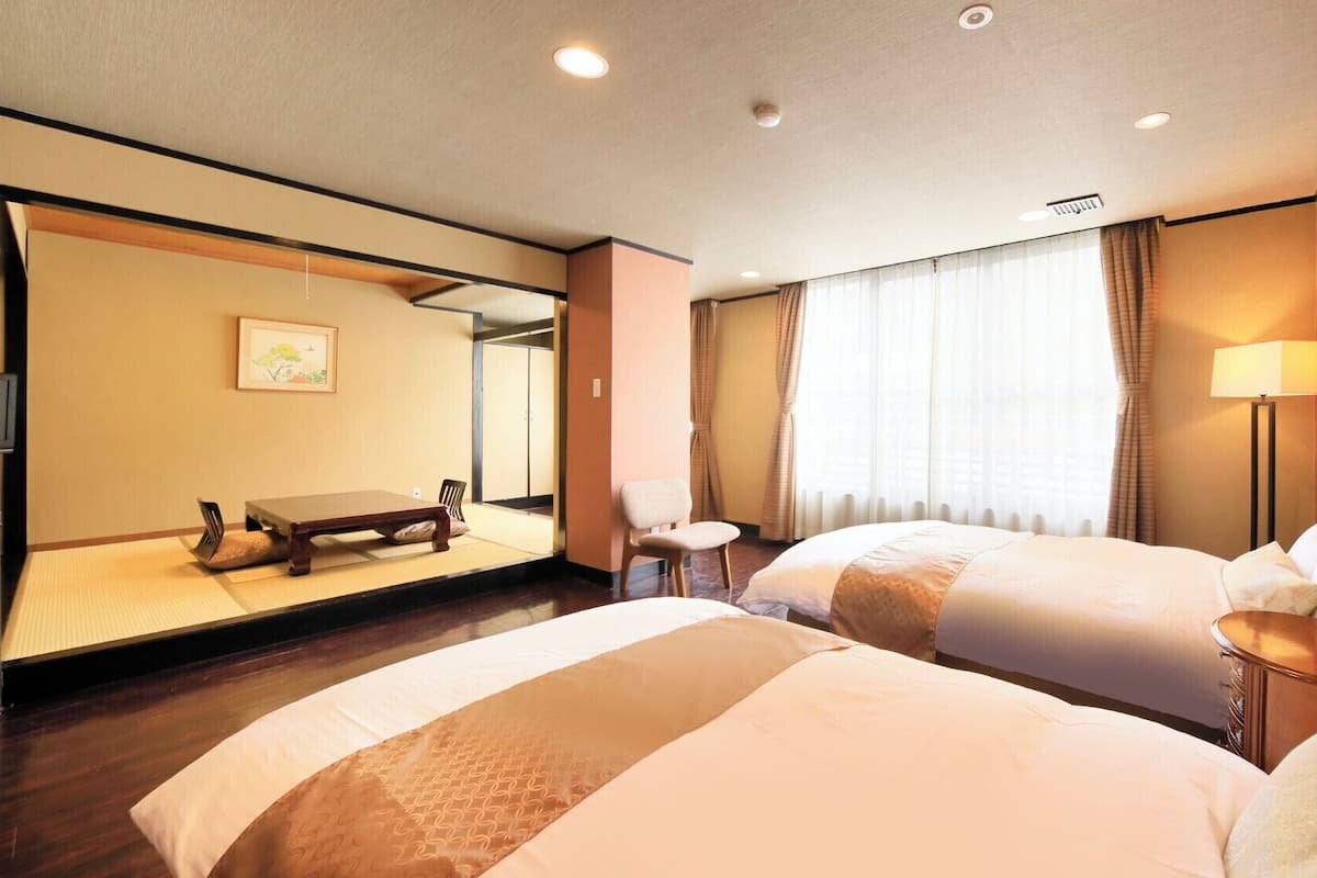 Habitación de hotel con dos camas, un rincón de descanso con una silla, una mesita, una lámpara y un ventanal grande con cortinas.