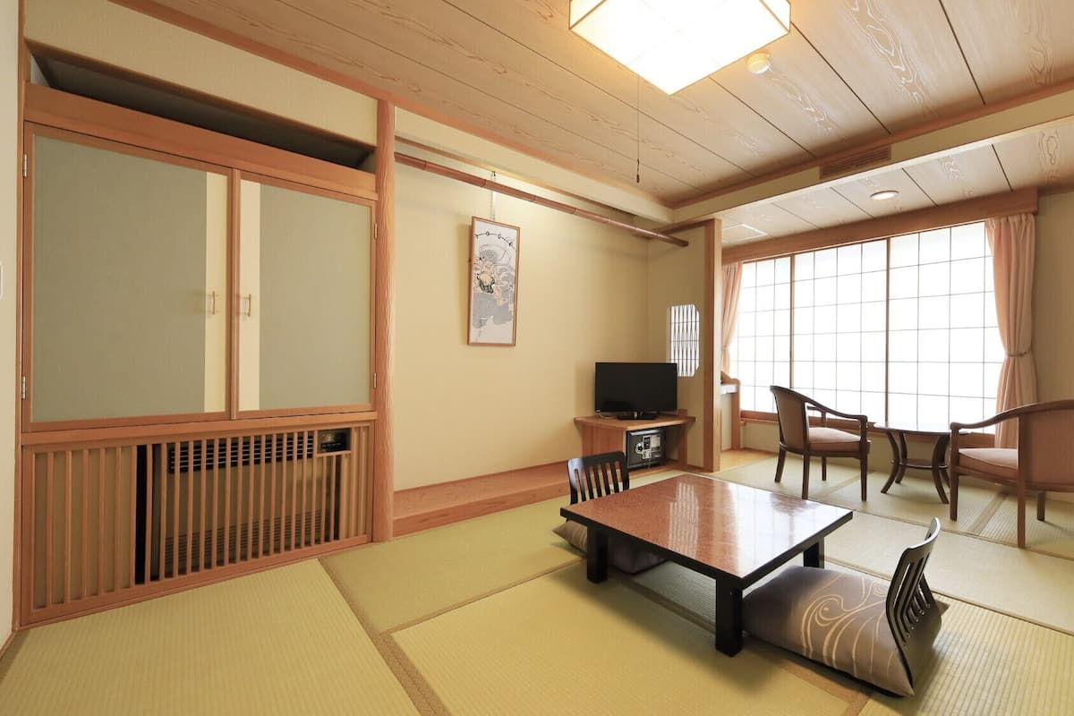 Una habitación japonesa tradicional con suelo de tatami, una mesa baja, sillas, un televisor y puertas correderas.