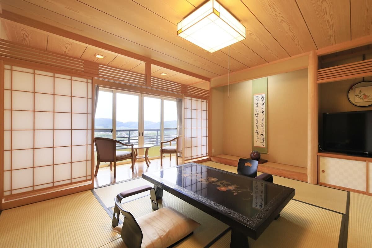 Una habitación japonesa tradicional con suelo de tatami, una mesa baja, sillas, un televisor y vistas a través de biombos shoji.