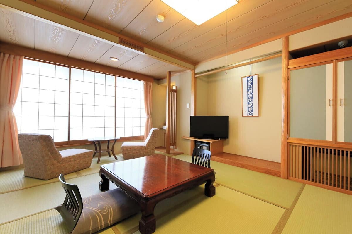 Una habitación japonesa tradicional con suelo de tatami, una mesa baja, sillas, un televisor y puertas correderas.
