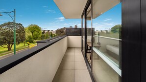 A balcony with a view of a park and buildings, a glass door, and a small table with a chair. - Whitehorse Apartment Hotel (Mont Albert)
