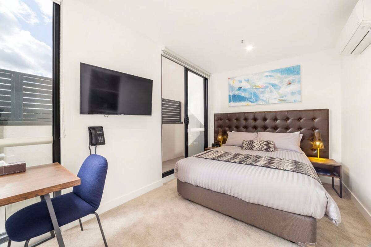 A bedroom with a large bed, a desk, a chair, a TV, and a painting on the wall.
