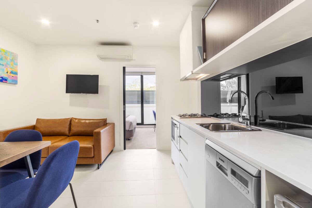 A modern kitchen with white cabinets, a built-in oven, and a dishwasher. There is a dining area with a wooden table and blue chairs. A brown leather sofa is in the living area, and a flat-screen TV is mounted on the wall.
