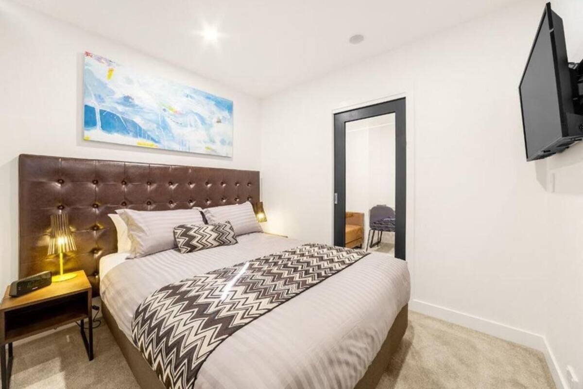 A bedroom with a tufted headboard, a bed with patterned bedding, a bedside lamp, a flat-screen TV mounted on the wall, and a painting above the bed.