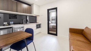A modern kitchen with white cabinets, a microwave, and a dishwasher. There is a blue chair and a wooden table. A brown sofa is visible in the foreground. - Whitehorse Apartment Hotel (Mont Albert)