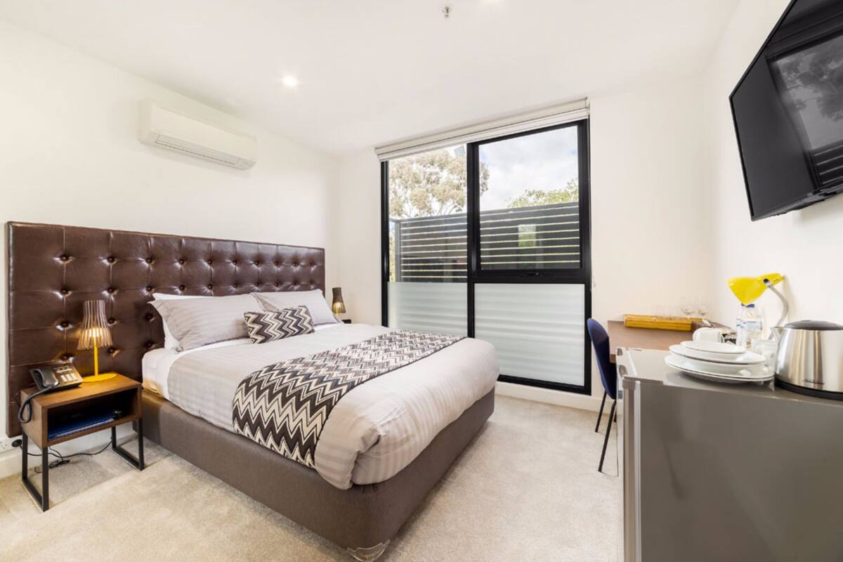 A modern bedroom with a tufted headboard, a large bed, a bedside table with a lamp, a TV mounted on the wall, and a window with blinds.