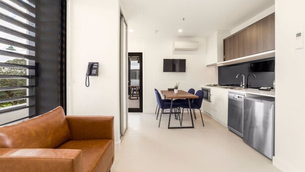 A modern kitchen with stainless steel appliances, a dining area with blue chairs, and a leather sofa. - Whitehorse Apartment Hotel (Mont Albert)