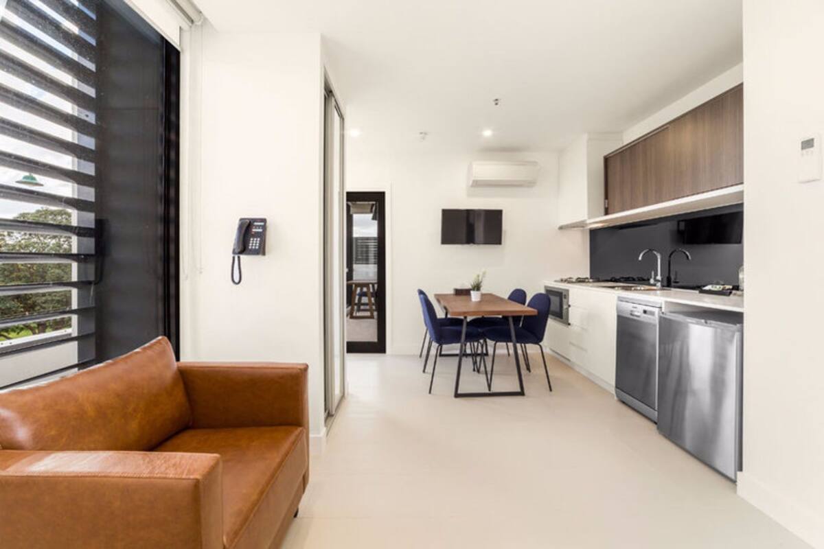 A modern kitchen with stainless steel appliances, a dining area with blue chairs, and a leather sofa.