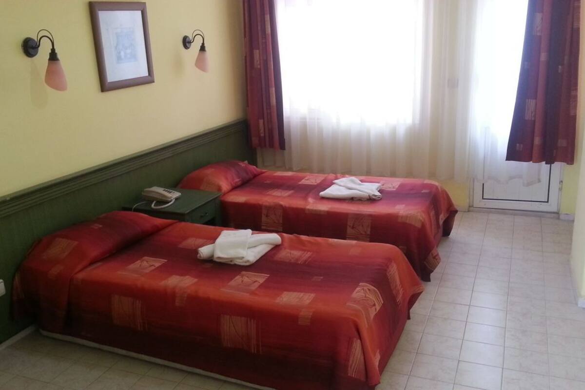 A hotel room with two beds, each with a red bedspread and white towels. There is a telephone on the nightstand, a framed picture on the wall, and a window with curtains.