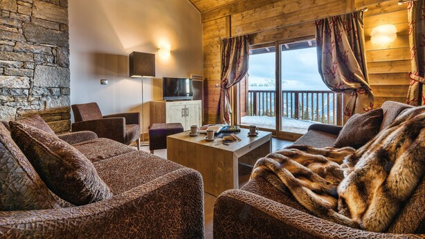 Interior acolhedor de uma cabana, com parede de pedra, teto de madeira, televisão, mesa de jantar e vista para o exterior.