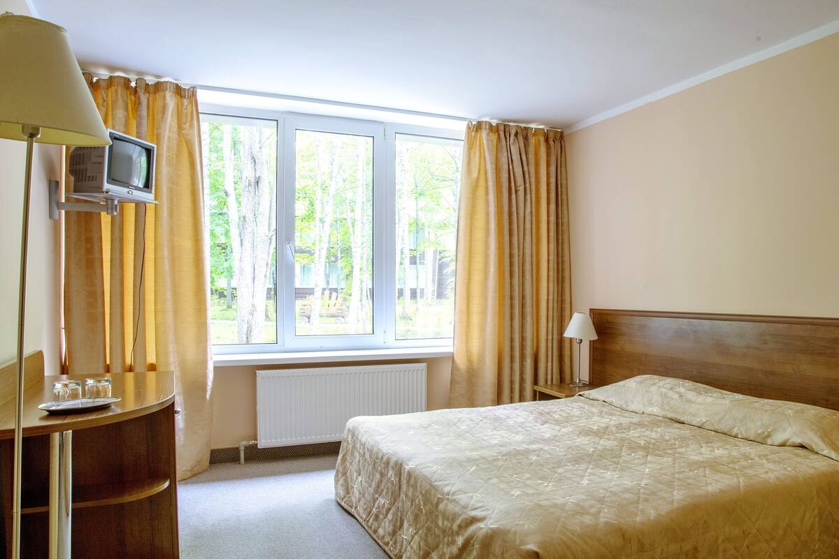 A hotel room with a bed, a desk with a lamp, a television, and a window with curtains.