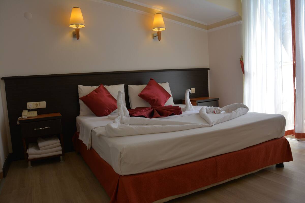 Habitación de hotel con una cama grande, dos lámparas de noche, una mesita de noche y una ventana con cortinas.
