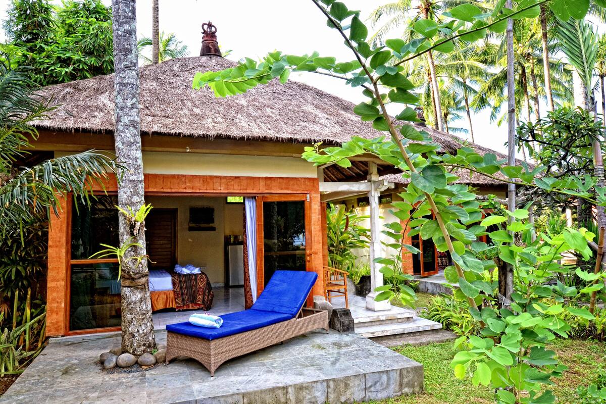 A thatched-roof bungalow with a lounge chair and a small table, surrounded by lush greenery.