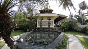 Une piscine bordée de pierres, entourée de plantes tropicales et un bâtiment doté d’un toit de chaume.
