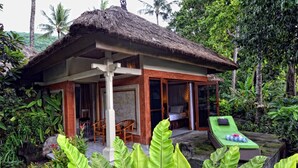 Un bungalow au toit de chaume, doté d’une véranda, d’un espace détente avec une table de massage, et entouré d’une végétation tropicale luxuriante.