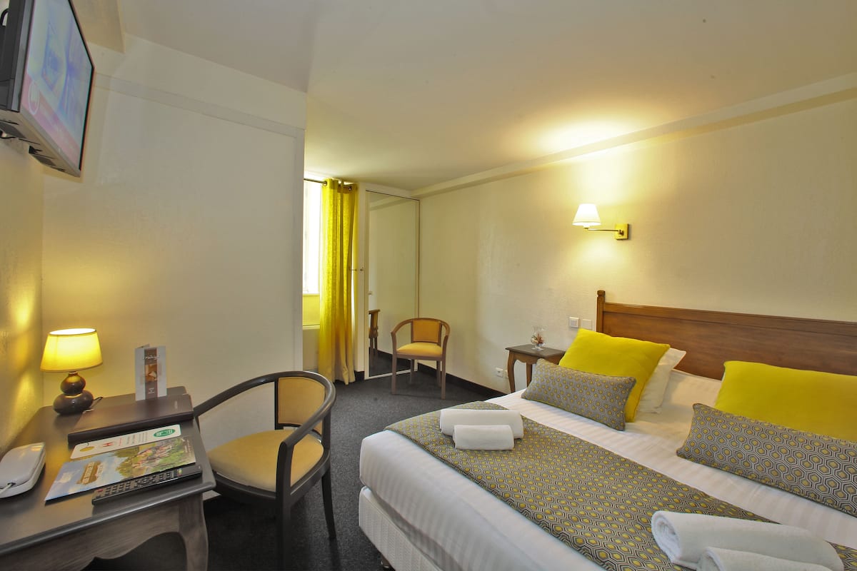 Una habitación de hotel con cama, escritorio con lámpara, silla y televisión en la pared.