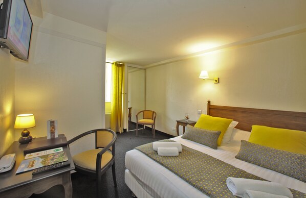 Una habitación de hotel con cama, escritorio con lámpara, silla y televisión en la pared.