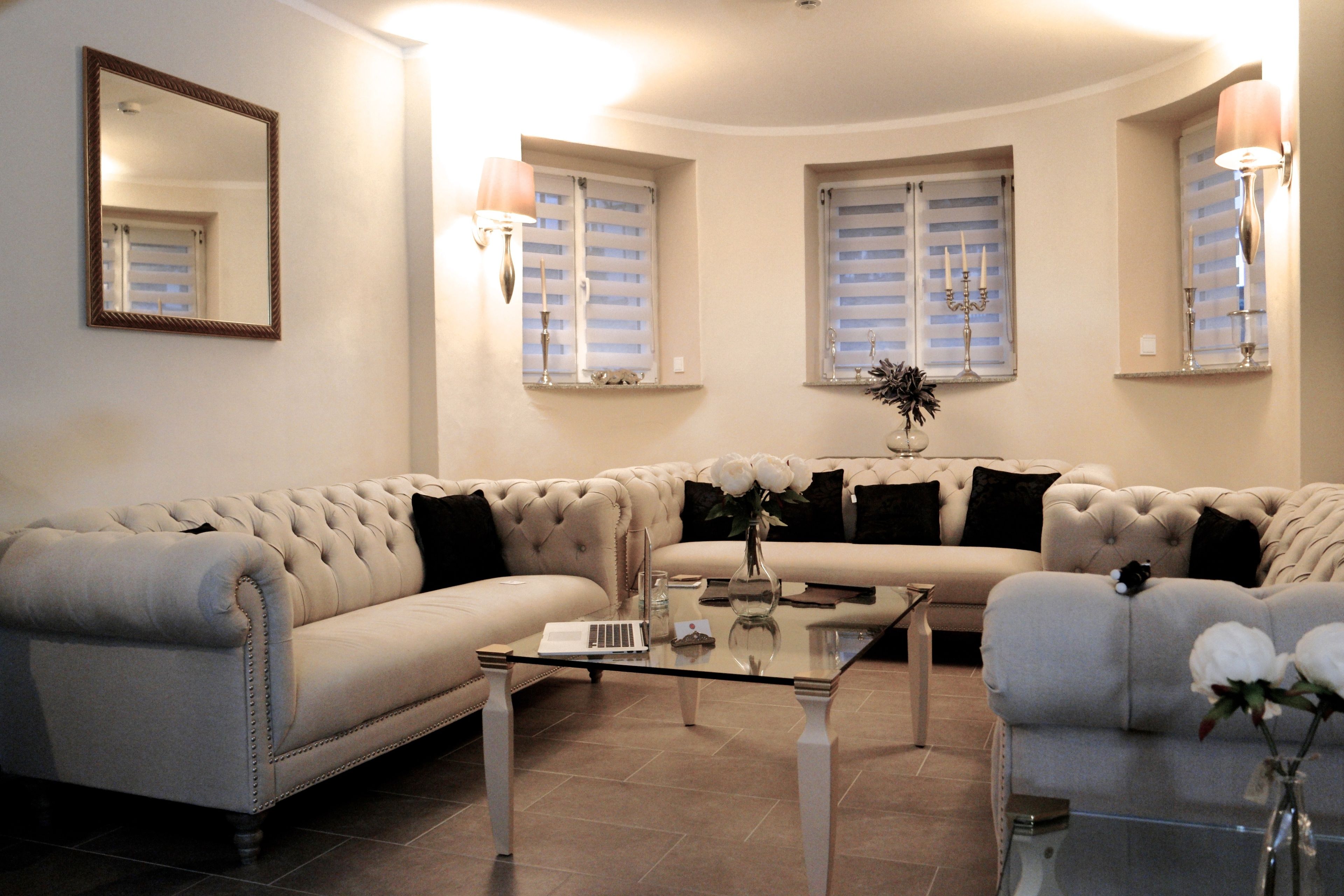 Una sala de estar con un sofá color beige, una mesa de centro de vidrio y un sillón blanco. Hay dos ventanas con persianas y lámparas de pared.
